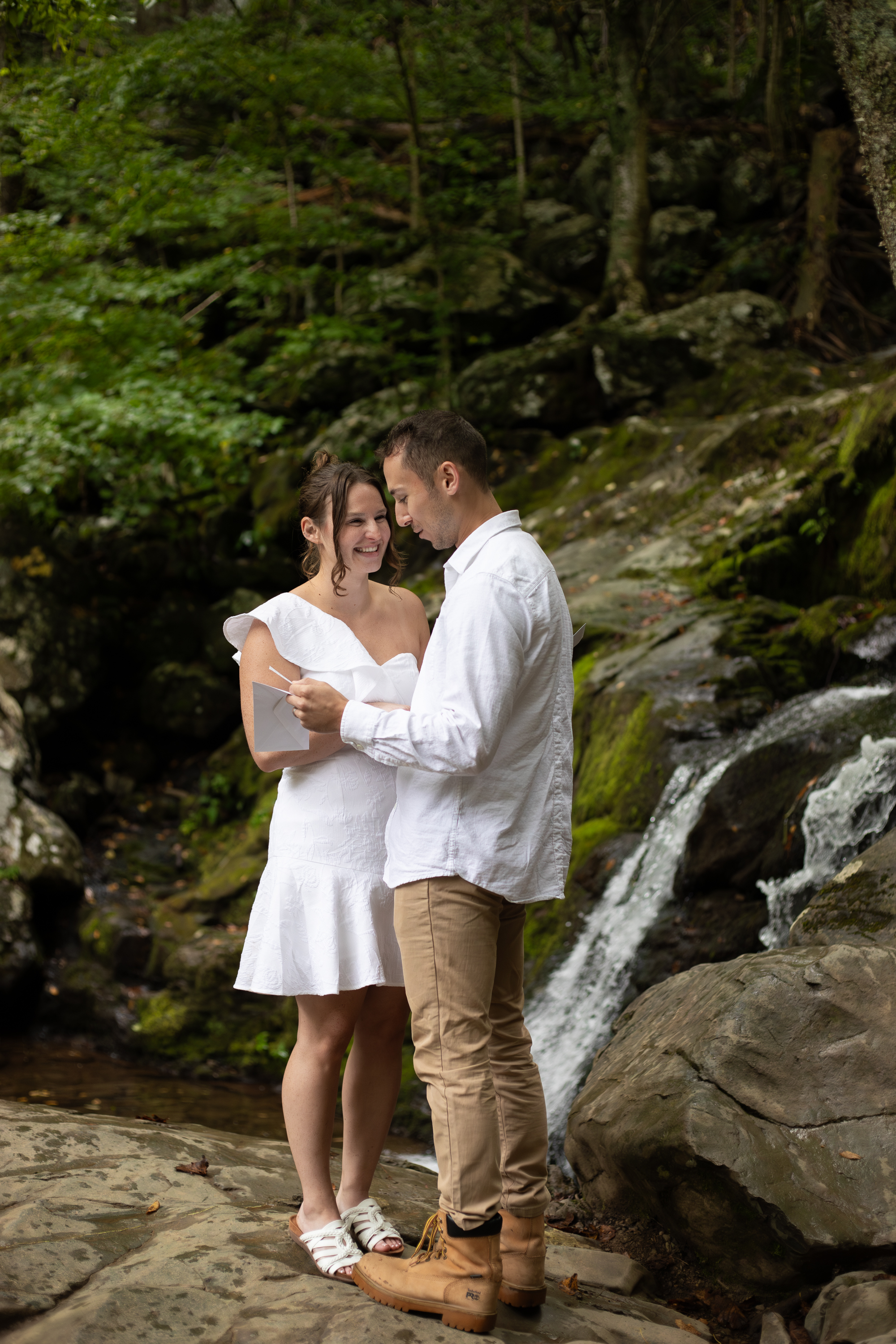 Couple exchanging vows at a Shenandoah waterfall, bride laughing while reading vows, groom smiling, moss-covered rocks and green forest behind them