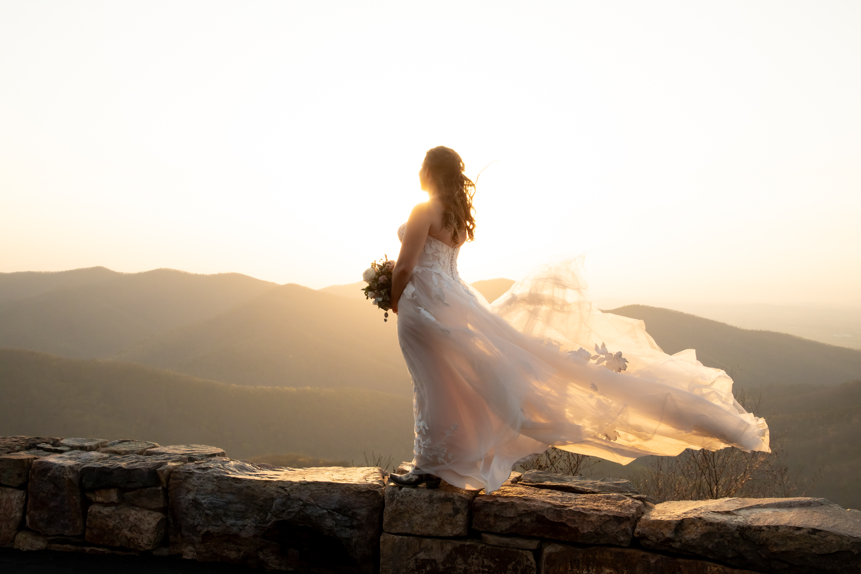 Bride in flowing white gown standing on mountain overlook at golden hour, dress billowing in the wind