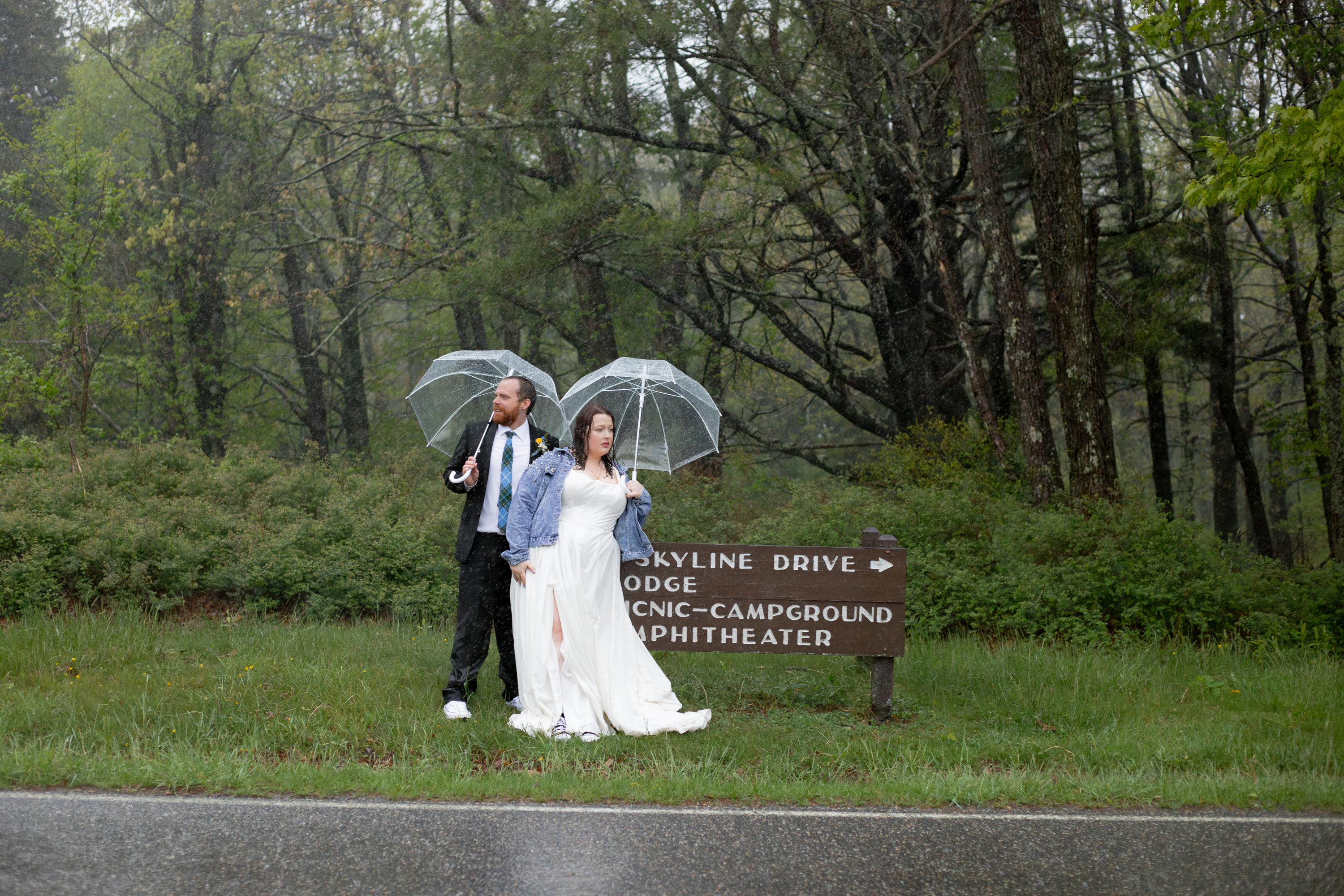 Couple standing next to a Skyline Drive sign in the rain, each holding a clear bubble umbrella, bride in a denim jacket and Converse sneakers