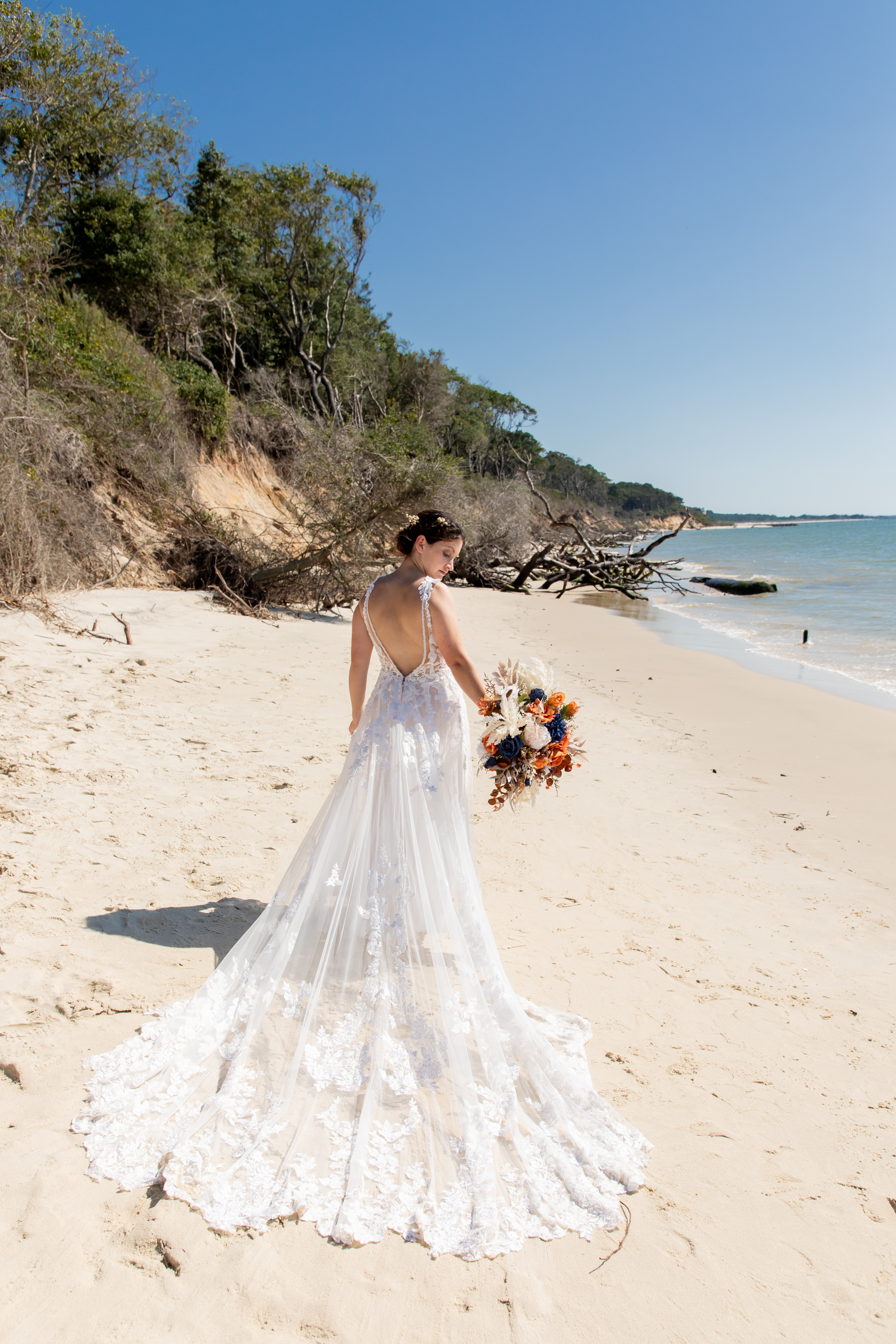 Bride in lace gown with long train on a Cape Charles beach with driftwood and calm bay water
