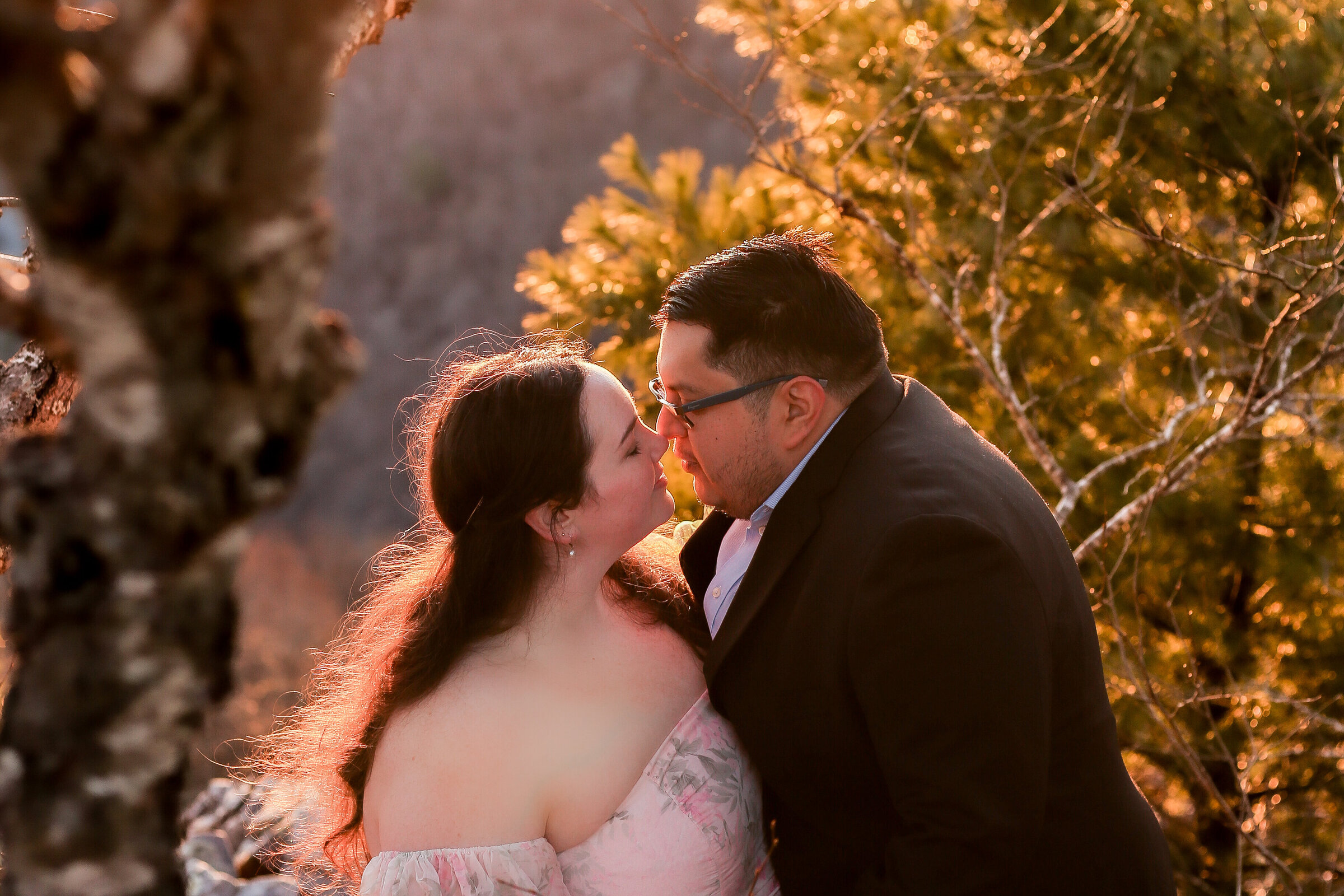Couple almost kissing in warm golden sunrise light among pine trees at a Shenandoah overlook, bride in a pink floral off-shoulder dress