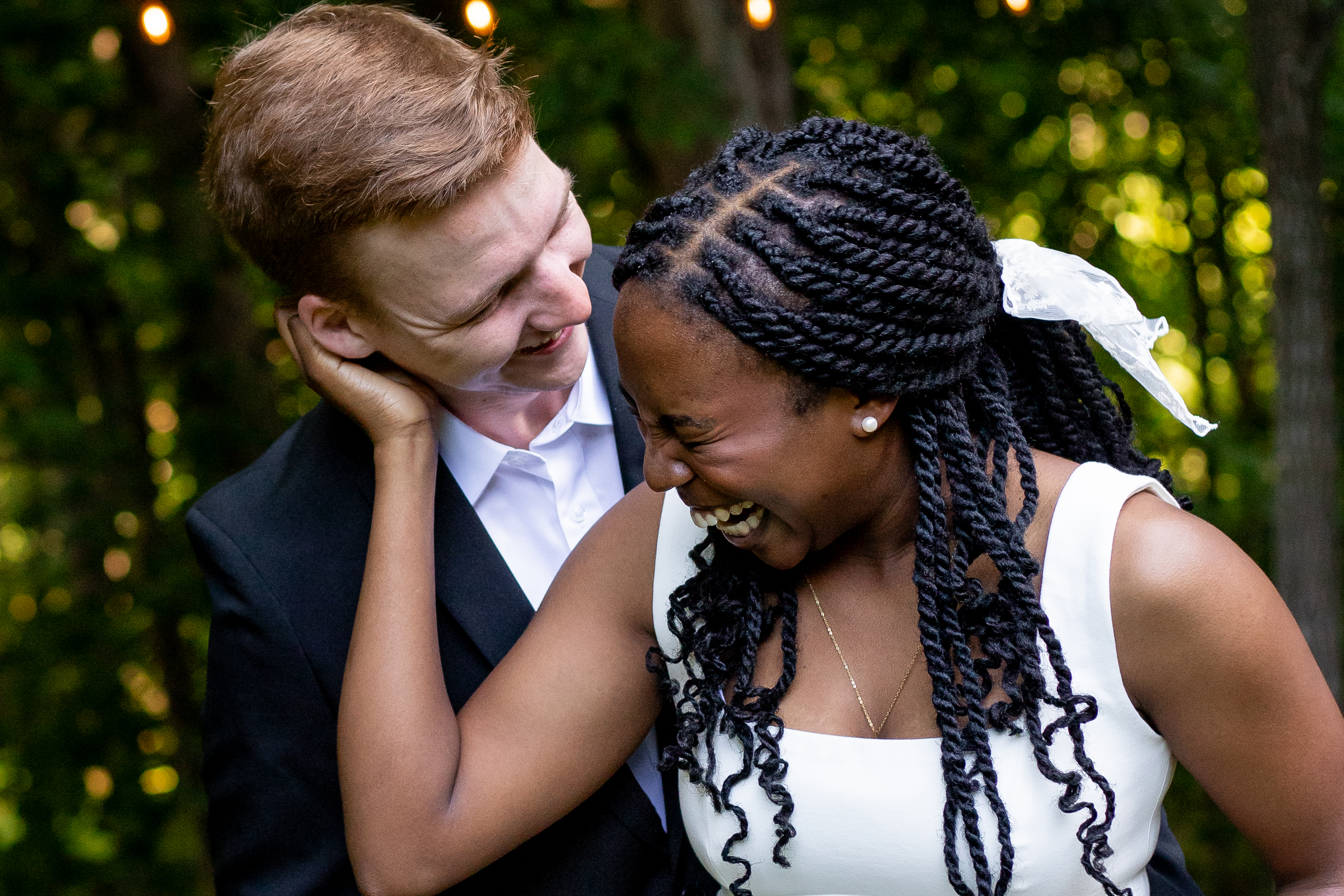 Bride laughing joyfully as groom leans in close, string lights and lush green trees behind them