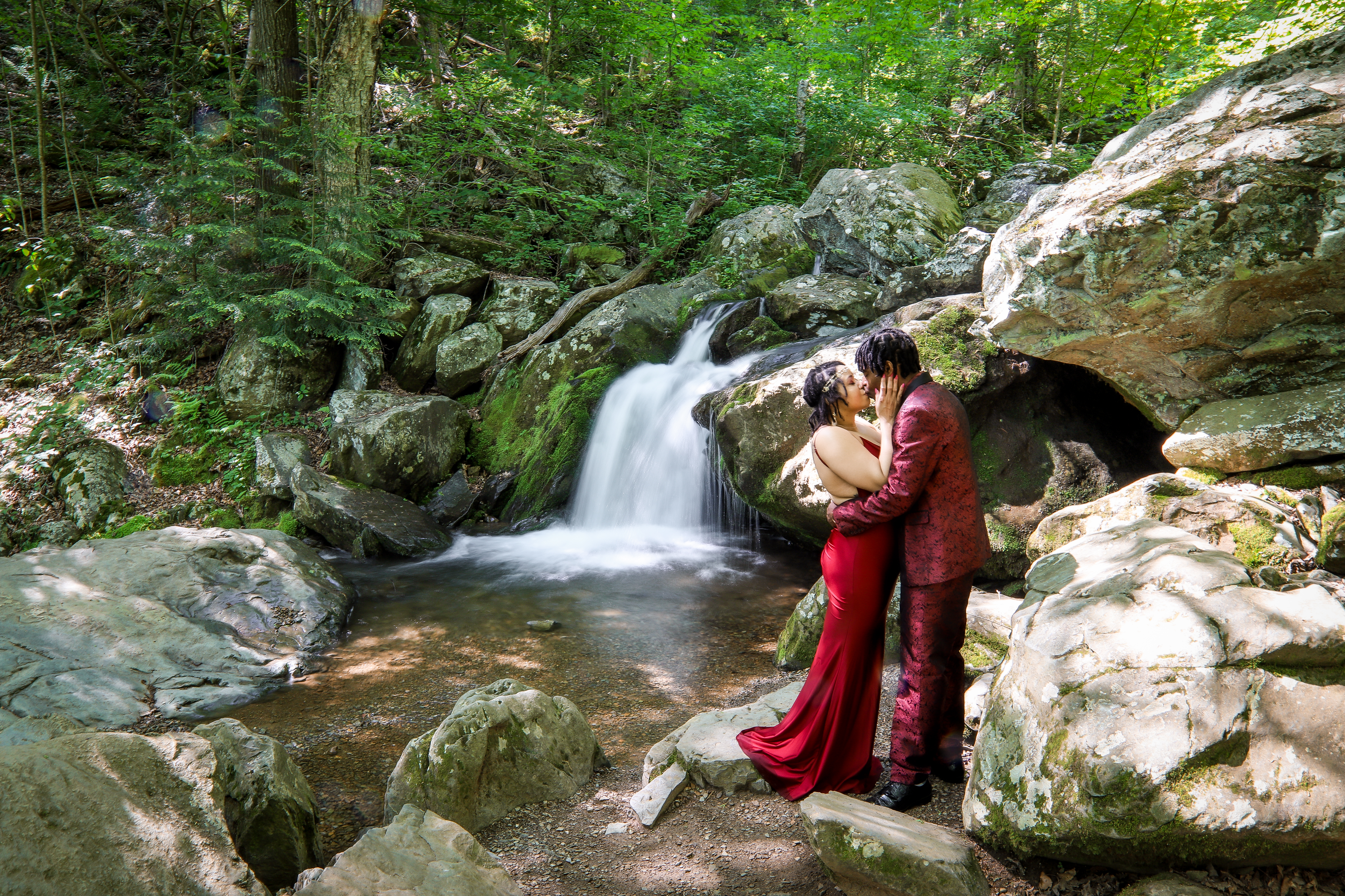 Couple in red kissing at a waterfall surrounded by lush green summer forest in Shenandoah National Park