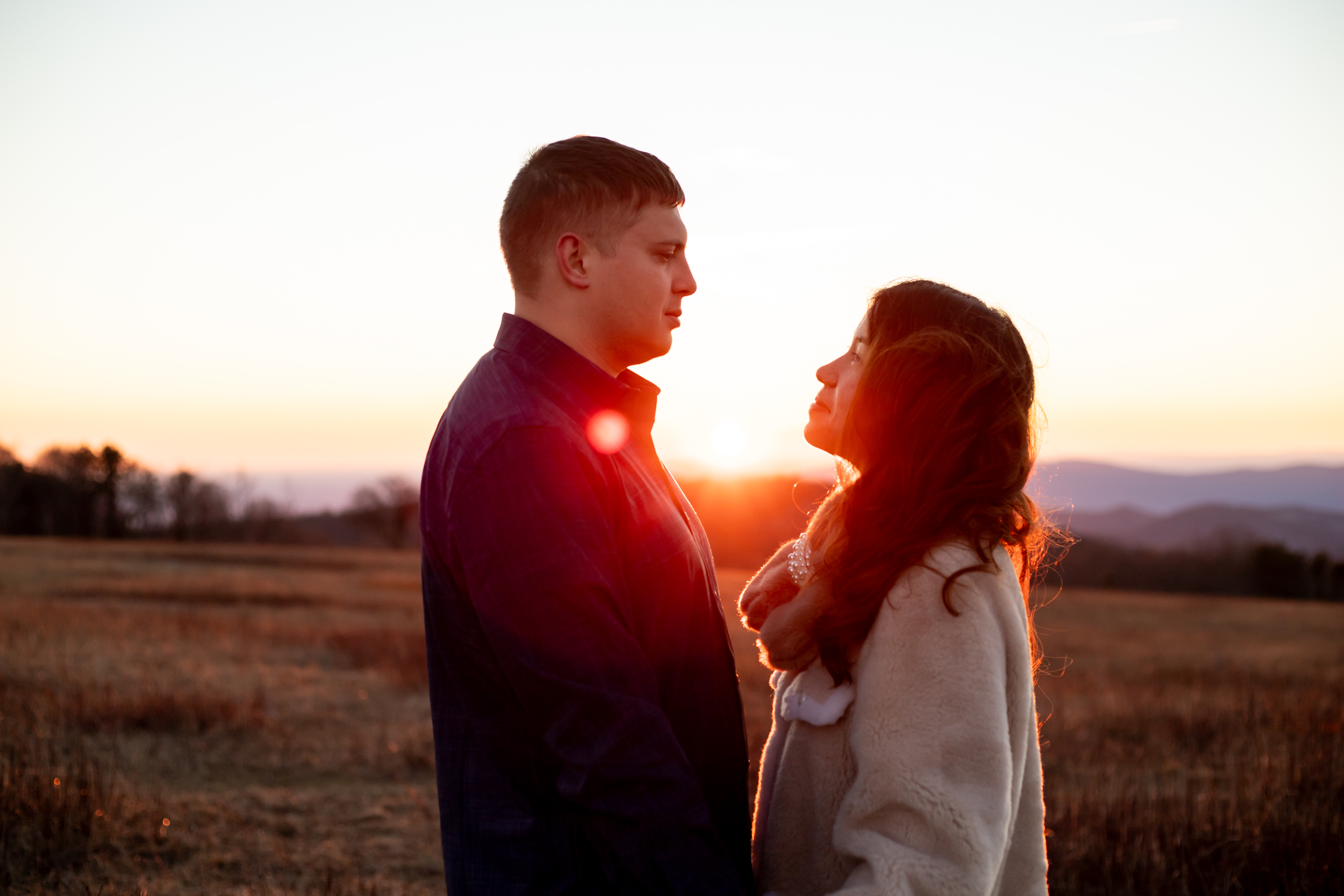 Couple facing each other at golden hour sunset in the open meadows of Big Meadows, Shenandoah National Park, woman in a white wrap coat with mountains glowing behind them