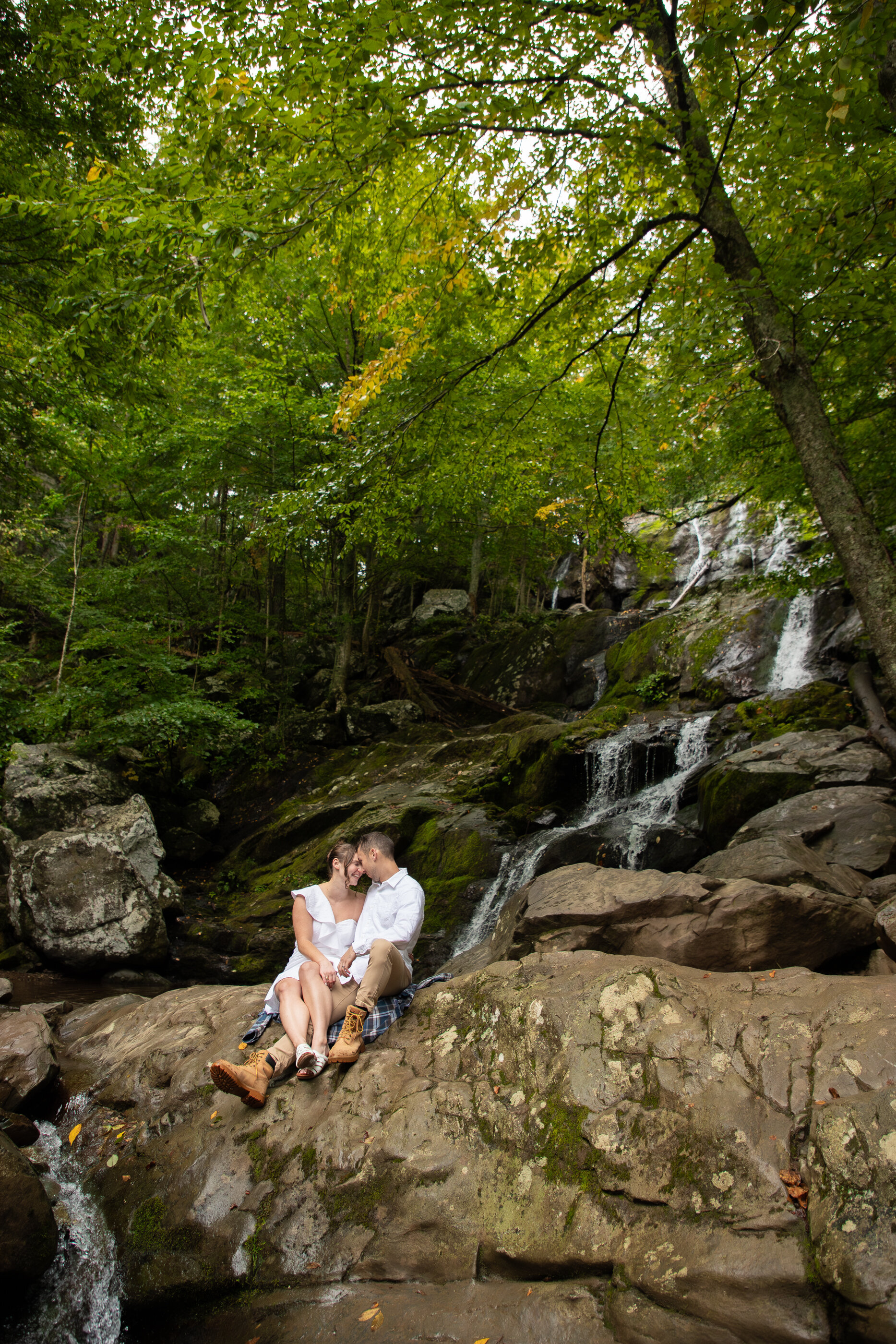 Couple sitting on mossy rocks at the base of Dark Hollow Falls in Shenandoah, laughing together beneath a lush green forest canopy
