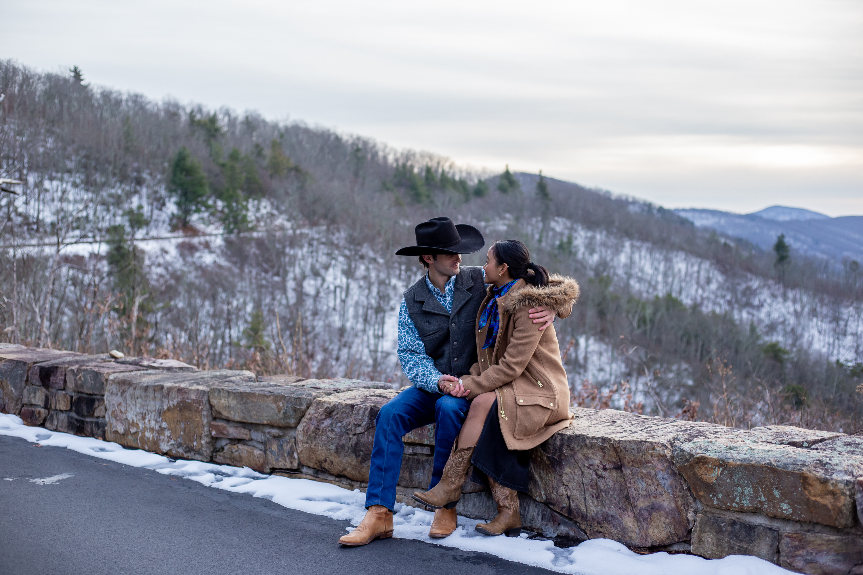 Couple sitting close together on a stone wall along Skyline Drive in winter with snow-covered mountains behind them