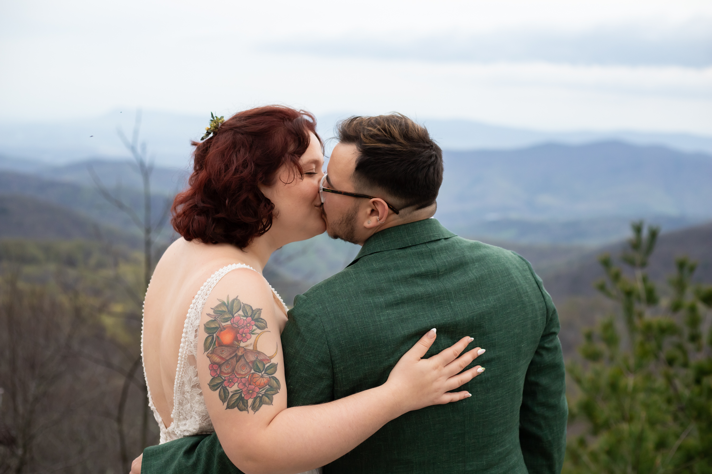Couple kissing at a Shenandoah overlook in spring, Blue Ridge mountains layering into the distance behind them, bride in a lace dress with a floral tattoo sleeve, groom in a forest green suit